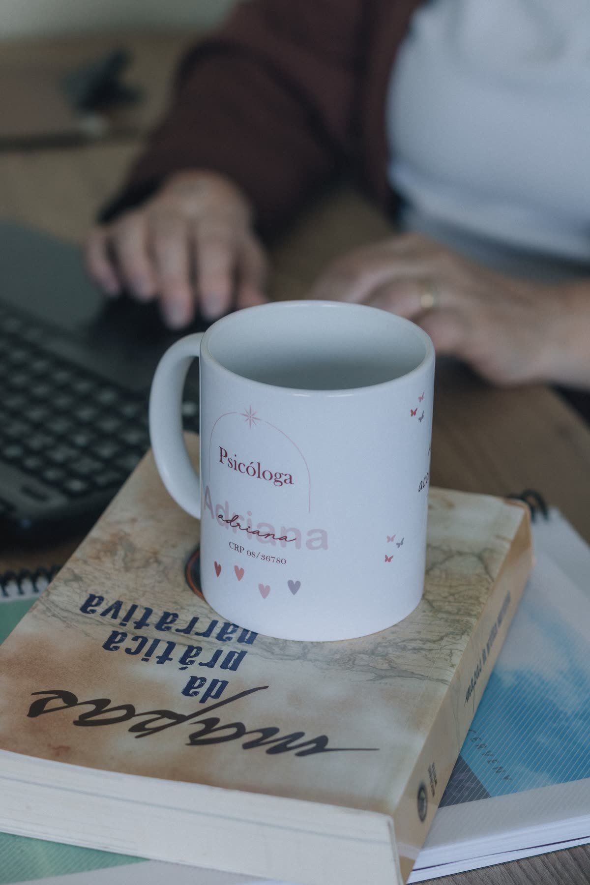 Taza de psicóloga sobre un libro de práctica narrativa junto a una laptop, representando el trabajo detrás de una clínica virtual
