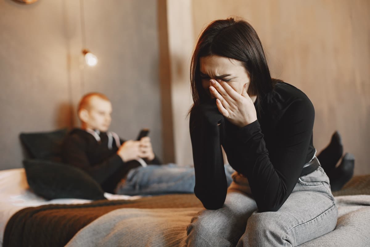 Mujer llorando en primer plano mientras su pareja está distraída al fondo, representando el desgaste emocional en la relación